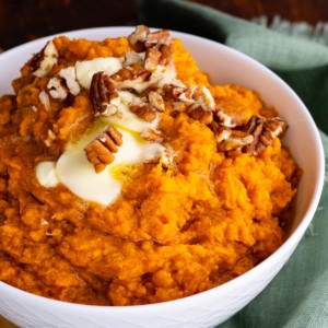 Mashed sweet potatoes in a white bowl with melted butter on top and chopped pecans. A teal tea towel to the side.