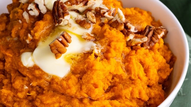Mashed sweet potatoes in a white bowl with melted butter on top and chopped pecans. A teal tea towel to the side.