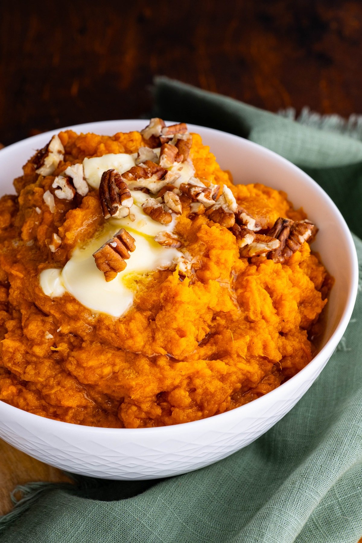 Mashed sweet potatoes in a white bowl with melted butter on top and chopped pecans. A teal tea towel to the side.