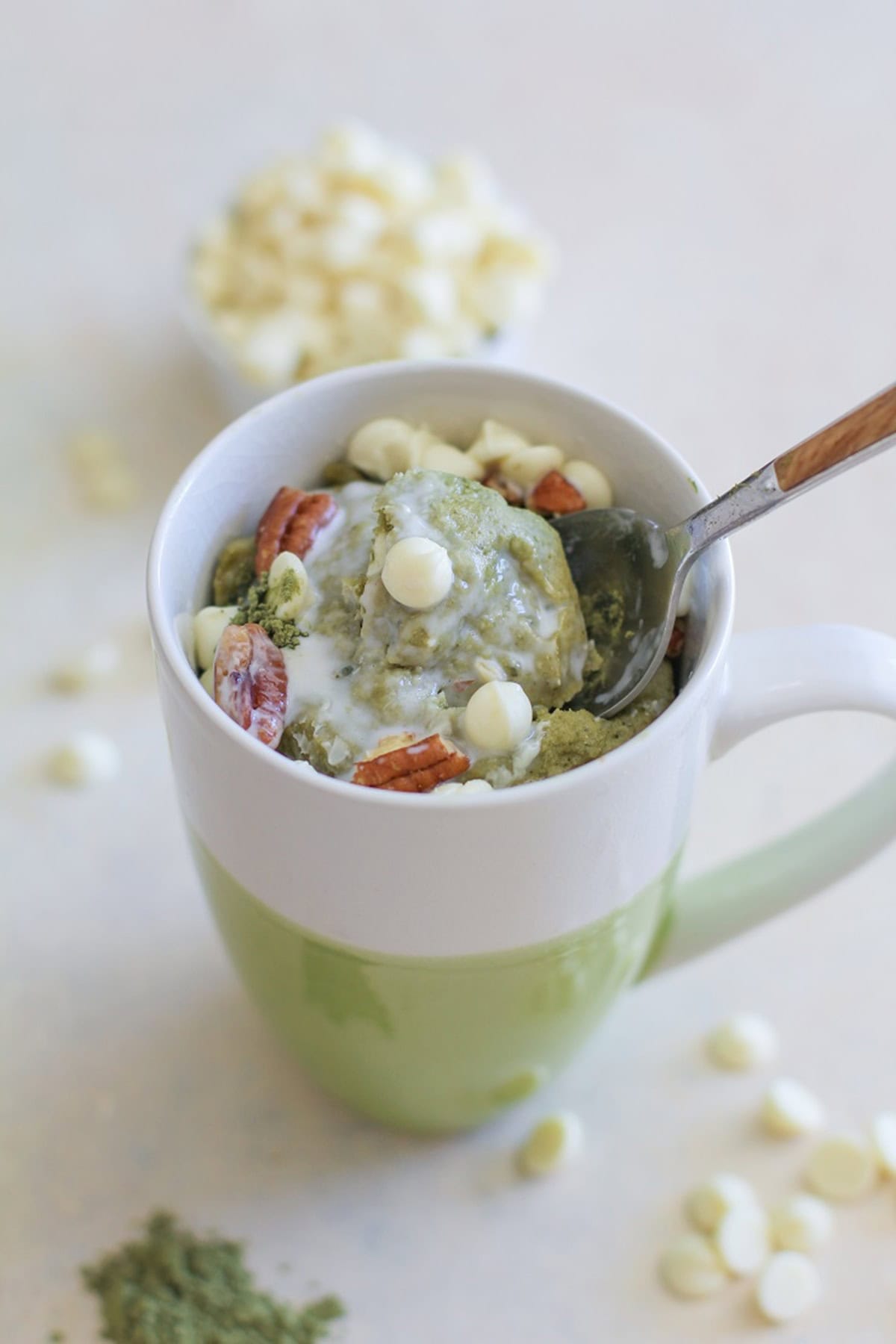 Matcha mug cake with a spoon taking a bite out of the center. white chocolate chips and pecans sprinkled on top of the cake.