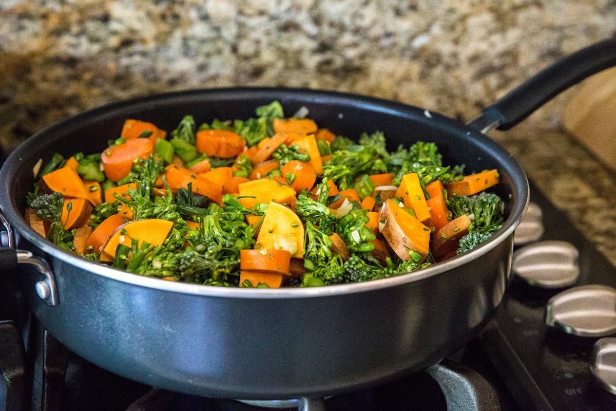 Raw sweet potatoes, broccoli, and onion cooking in a large skillet on the stove top.