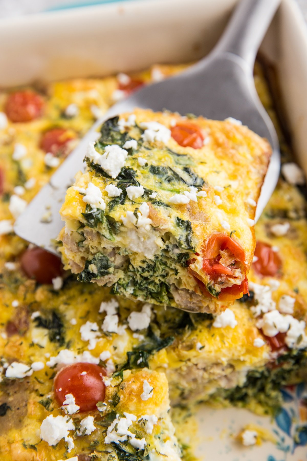 Healthy Breakfast Casserole with sausage, kale, cheese, and tomatoes in a large casserole dish with a spatula lifting out a piece.