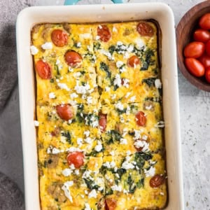 Casserole dish with a Mediterranean breakfast casserole inside. A napkin to the side and a bowl of fresh cherry tomatoes