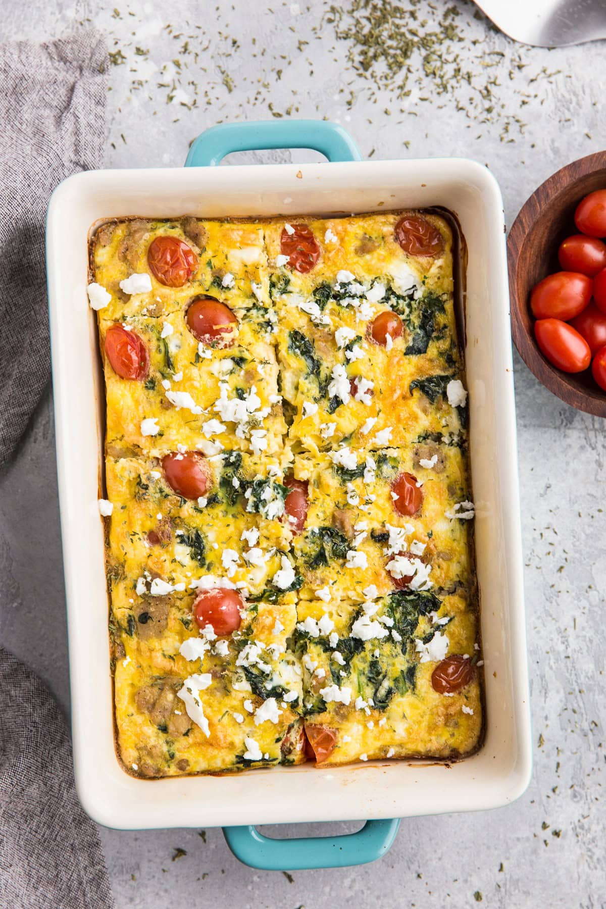 Casserole dish with a Mediterranean breakfast casserole inside. A napkin to the side and a bowl of fresh cherry tomatoes