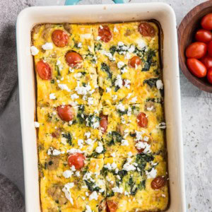 Mediterranean Breakfast Casserole fresh out of the oven in a casserole dish with dried herbs sprinkled all around, a napkin, and a bowl of fresh cherry tomatoes.