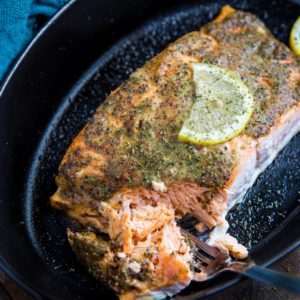 Large salmon filet in a baking dish with a coat of mustard and seasonings on top for a baked mustard salmon recipe. A fork taking bites at the fish to show how flaky and moist it is. A slice of lemon on top of the fish.