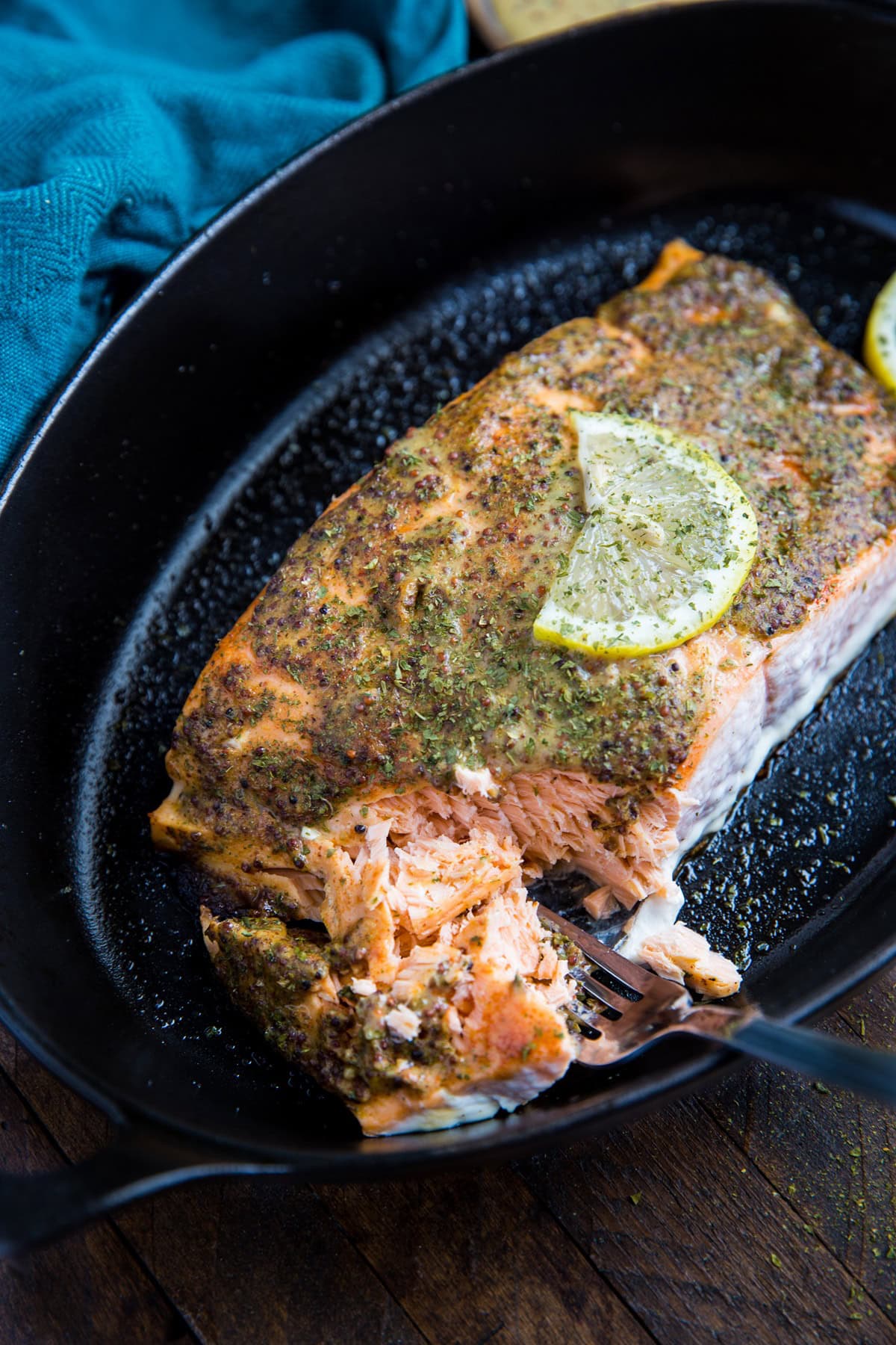 Large salmon filet in a baking dish with a coat of mustard and seasonings on top for a baked mustard salmon recipe. A fork taking bites at the fish to show how flaky and moist it is. A slice of lemon on top of the fish.