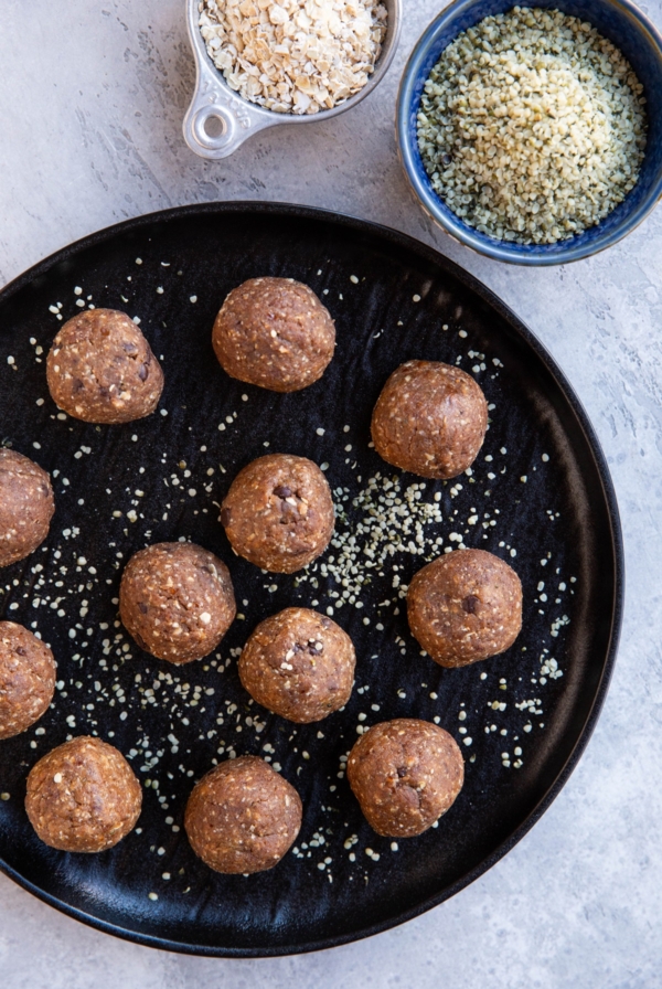 Black plate full of energy balls sprinkled with hemp seeds. A bowl and measuring cup of oats and hemp seeds to the side.