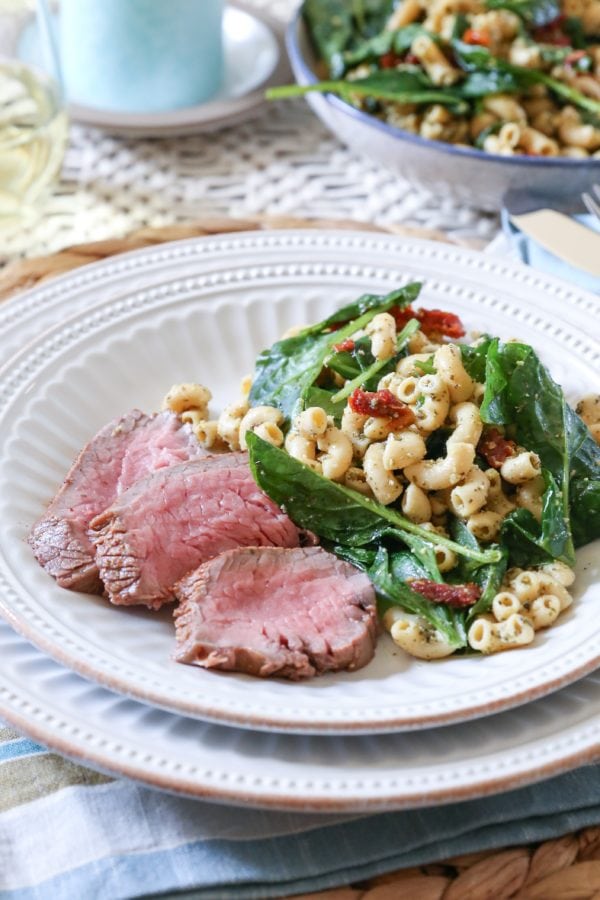 Slices of oven baked tri tip on a white plate with macaroni salad. A place mat and napkin underneath the plate and a glass of wine in the background.