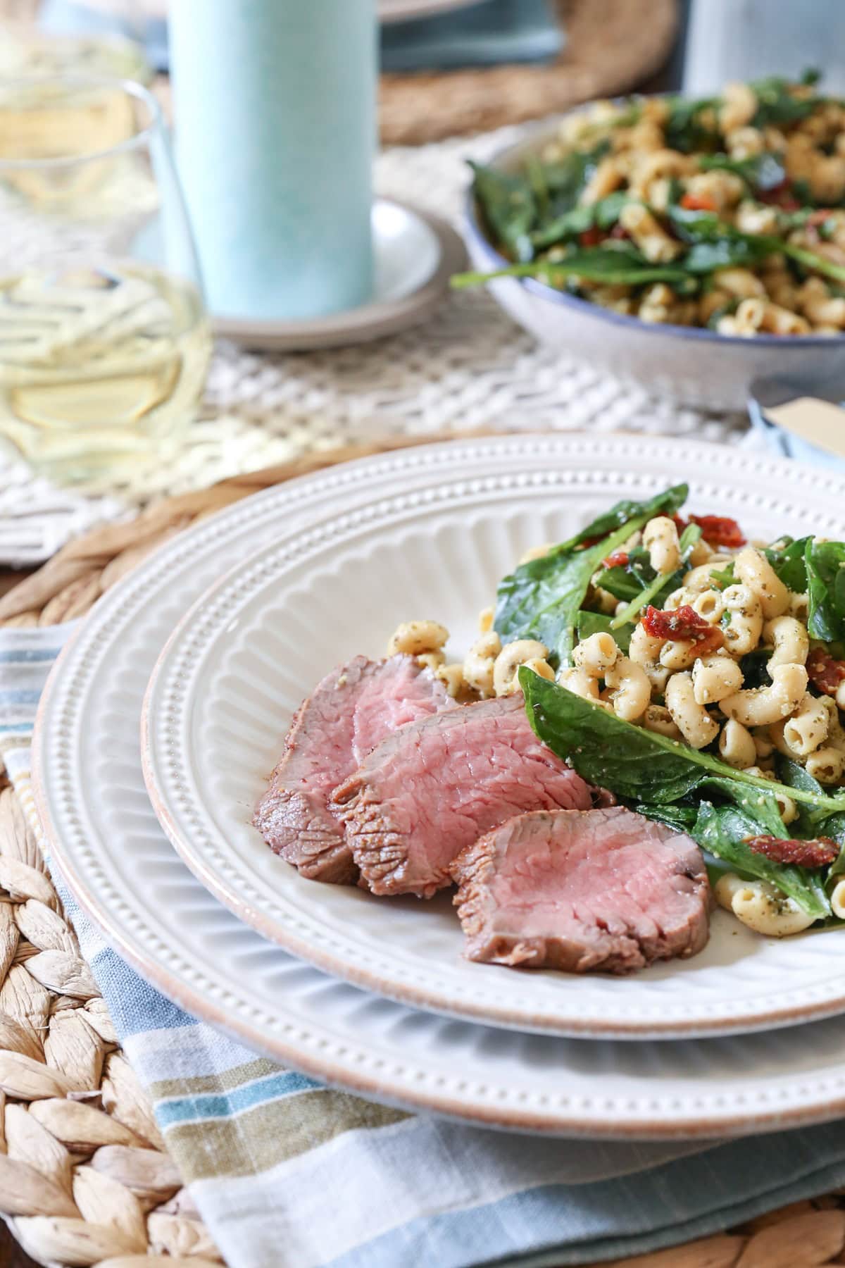 Tri tip slices on a white plate next to macaroni salad, ready to serve. Plate is on top of a placemat with a napkin and there's a bowl of macaroni salad in the background.