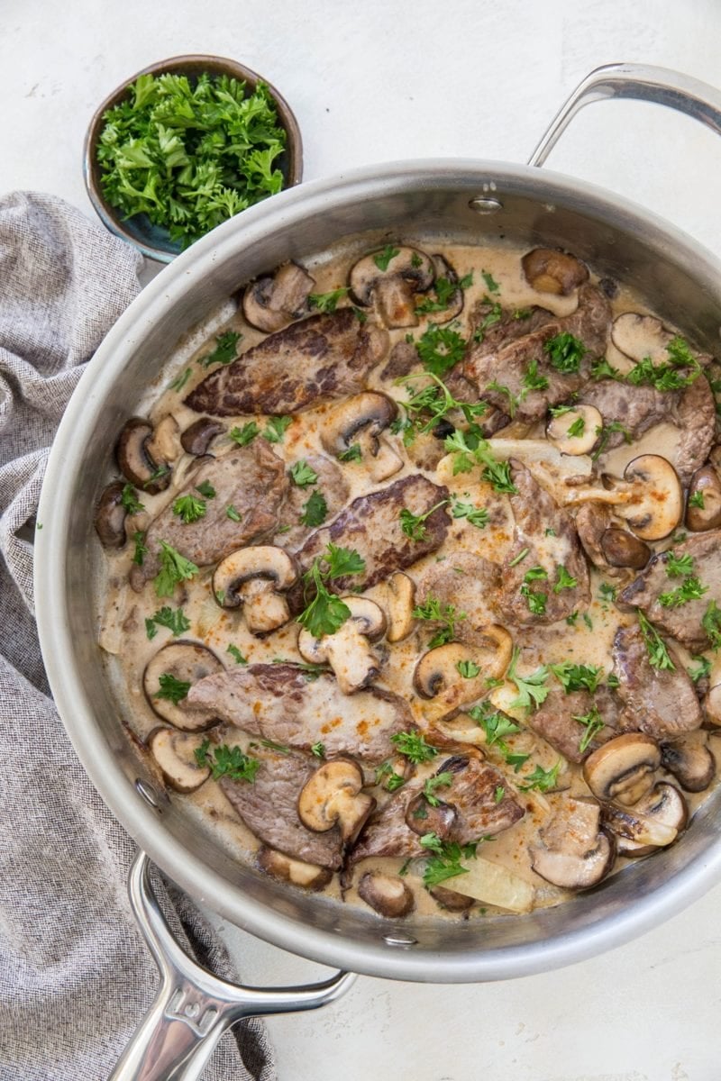 A stainless steel skillet filled with creamy paleo beef stroganoff, featuring sliced beef, mushrooms, onions, and a garnish of chopped parsley. A small bowl of fresh parsley and a gray cloth sit beside the skillet.