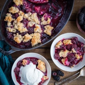 Paleo Blackberry Cobbler in two white bowls, one with a scoop of vanilla ice cream on top. The rest of the cobbler in a baking dish and fresh blackberries and spoons to the side.