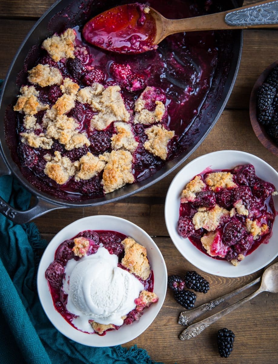 Paleo Blackberry Cobbler in two white bowls, one with a scoop of vanilla ice cream on top. The rest of the cobbler in a baking dish and fresh blackberries and spoons to the side.