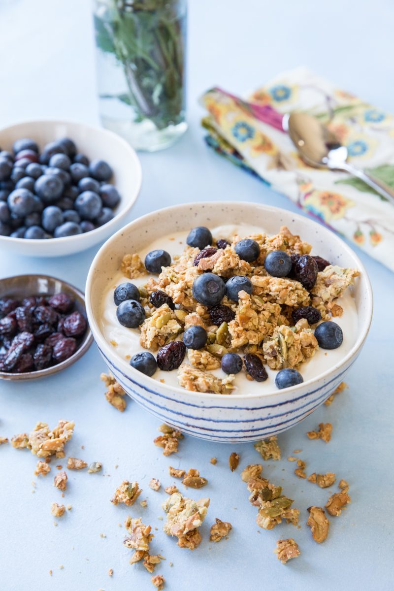white and blue striped bowl full of yogurt with paleo granola and fresh blueberries on top. Clusters of granola all around the bowl.