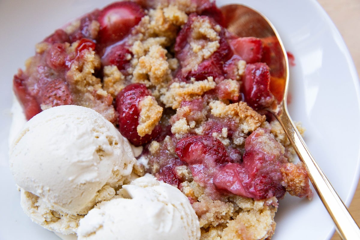 White bowl of strawberry crumble with scoops of vanilla ice cream.