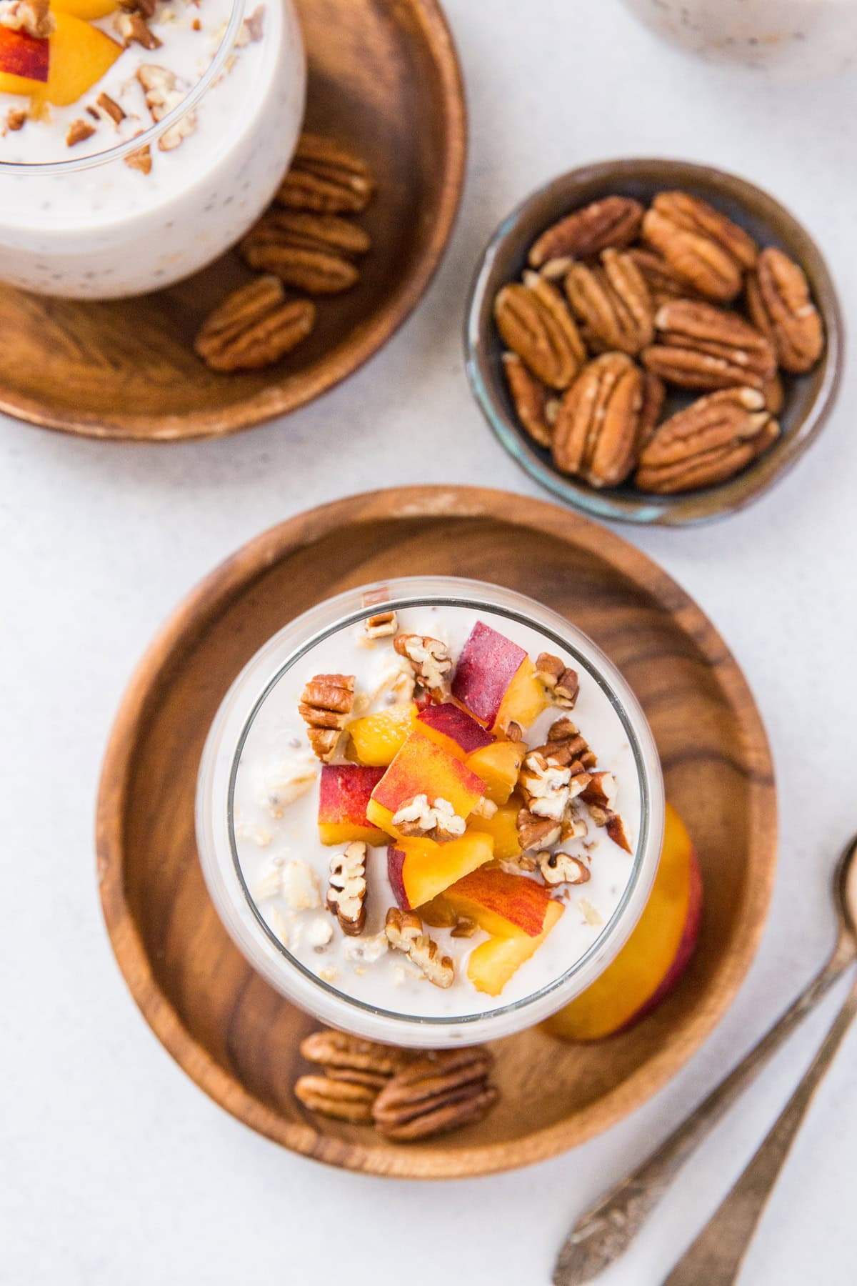 Peaches and cream overnight oats in wine glasses with chopped peaches and pecans on top. Spoons for serving.