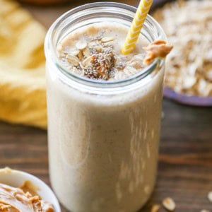 Banana Bread Oat Protein Smoothie in a glass with a yellow striped straw, ready to drink. Peanut butter, oats, and bananas in the background.