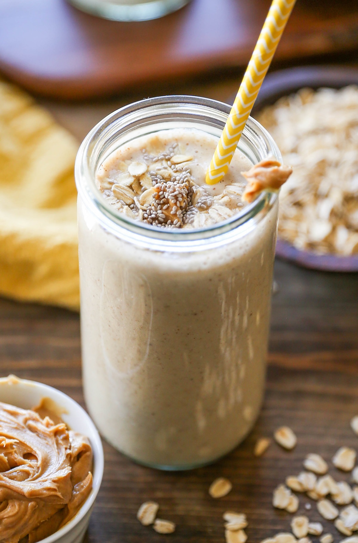 Banana Bread Oat Protein Smoothie in a glass with a yellow striped straw, ready to drink. Peanut butter, oats, and bananas in the background.