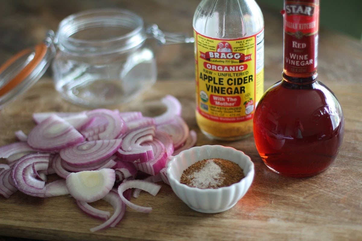 Wooden cutting board with sliced red onions a bowl of sugar and salt, red wine vinegar and apple cider vinegar and a open glass jar in the background. Ingredients to make pickled red onions.