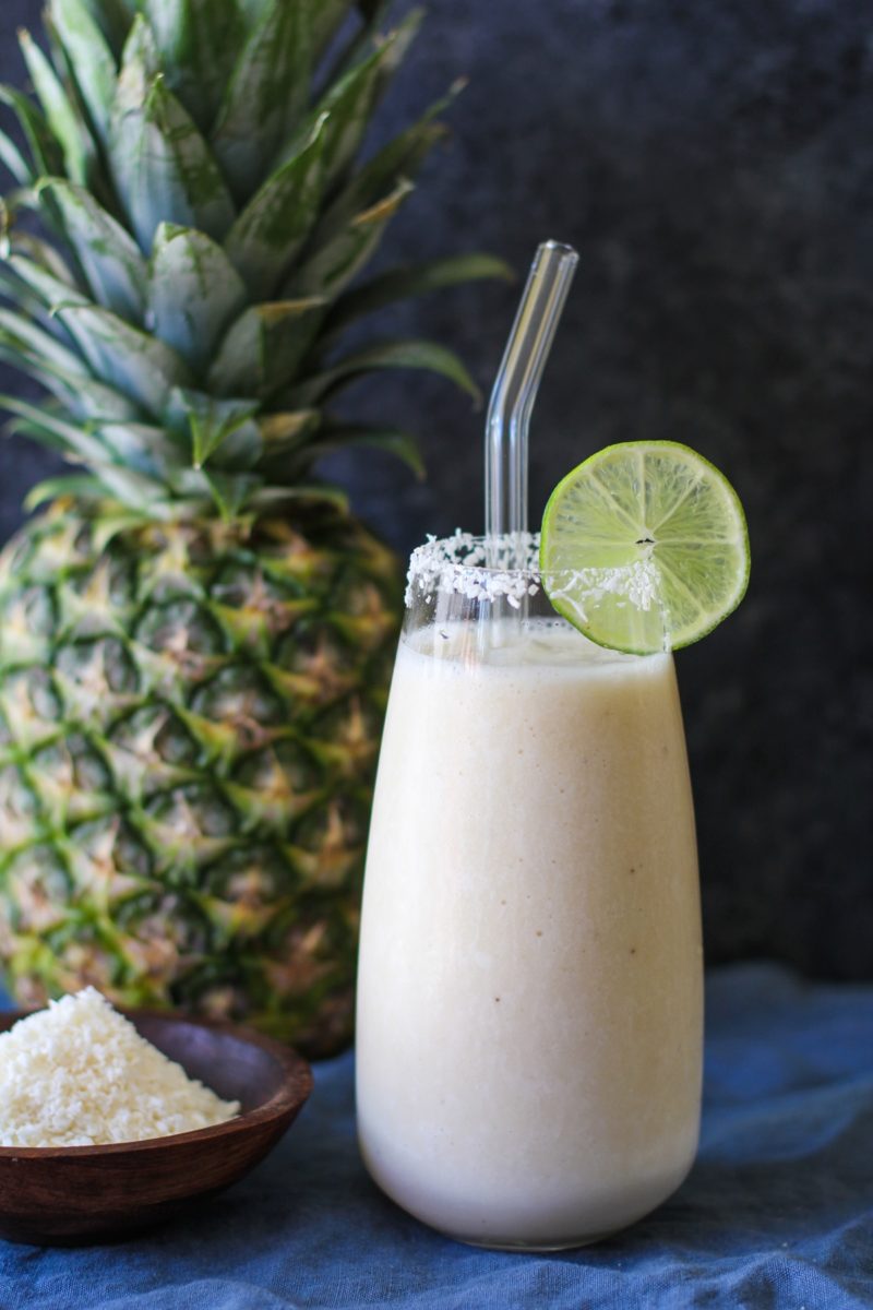 Pina colada smoothie in a glass with shredded coconut around the rim and a slice of lime with a glass straw, ready to drink.