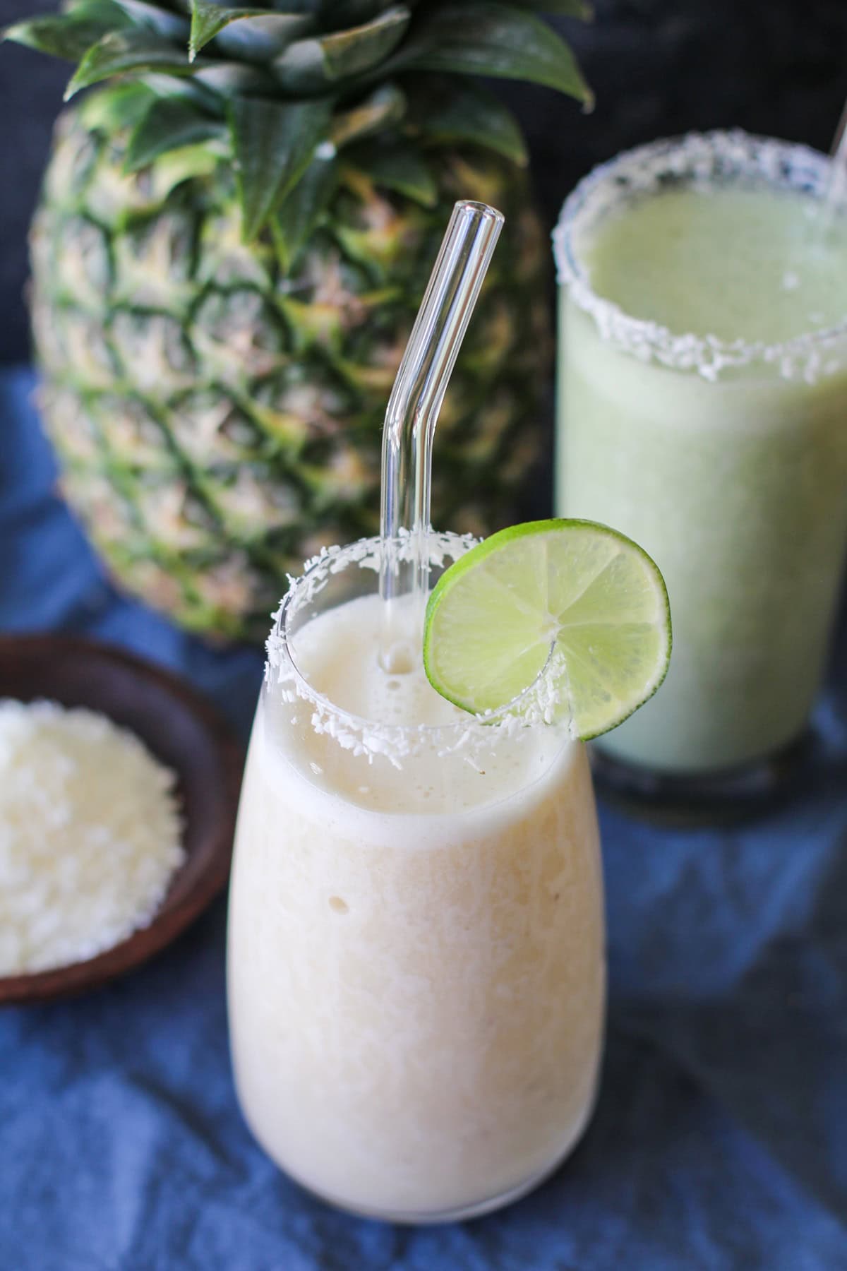 A creamy pina colada smoothie in a glass with a glass straw, garnished with a lime slice and a coconut rim. Another smoothie and a fresh pineapple rest in the background on a blue cloth.