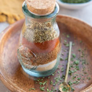 Jar full of seasonings with a cork on top and a small spoon to the side with dried parsley around the jar of seasonings and a small bowl of dried parsley in the background.