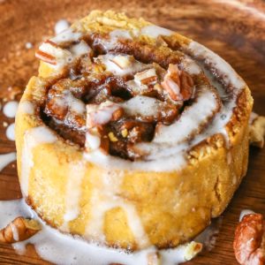 Large pumpkin cinnamon roll with pecans and glaze on a wooden plate, ready to eat.