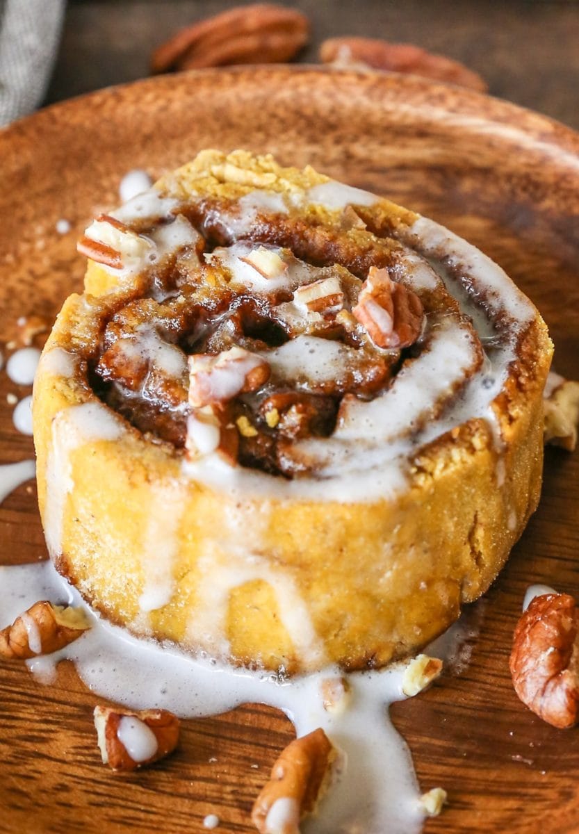 Large pumpkin cinnamon roll with pecans and glaze on a wooden plate, ready to eat.