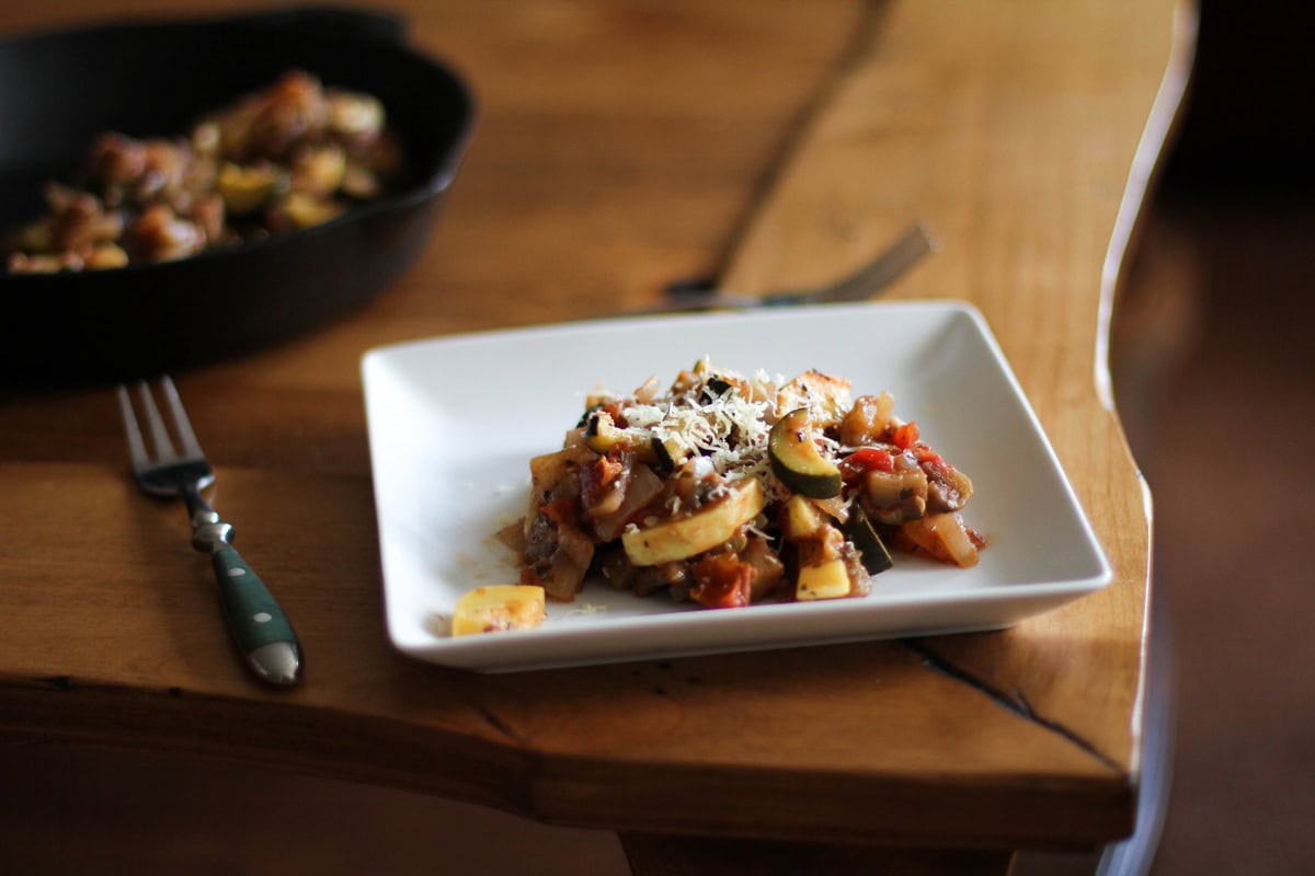 White plate of ratatouille on a table with a fork to the side and the rest of the skillet of ratatouille in the background.