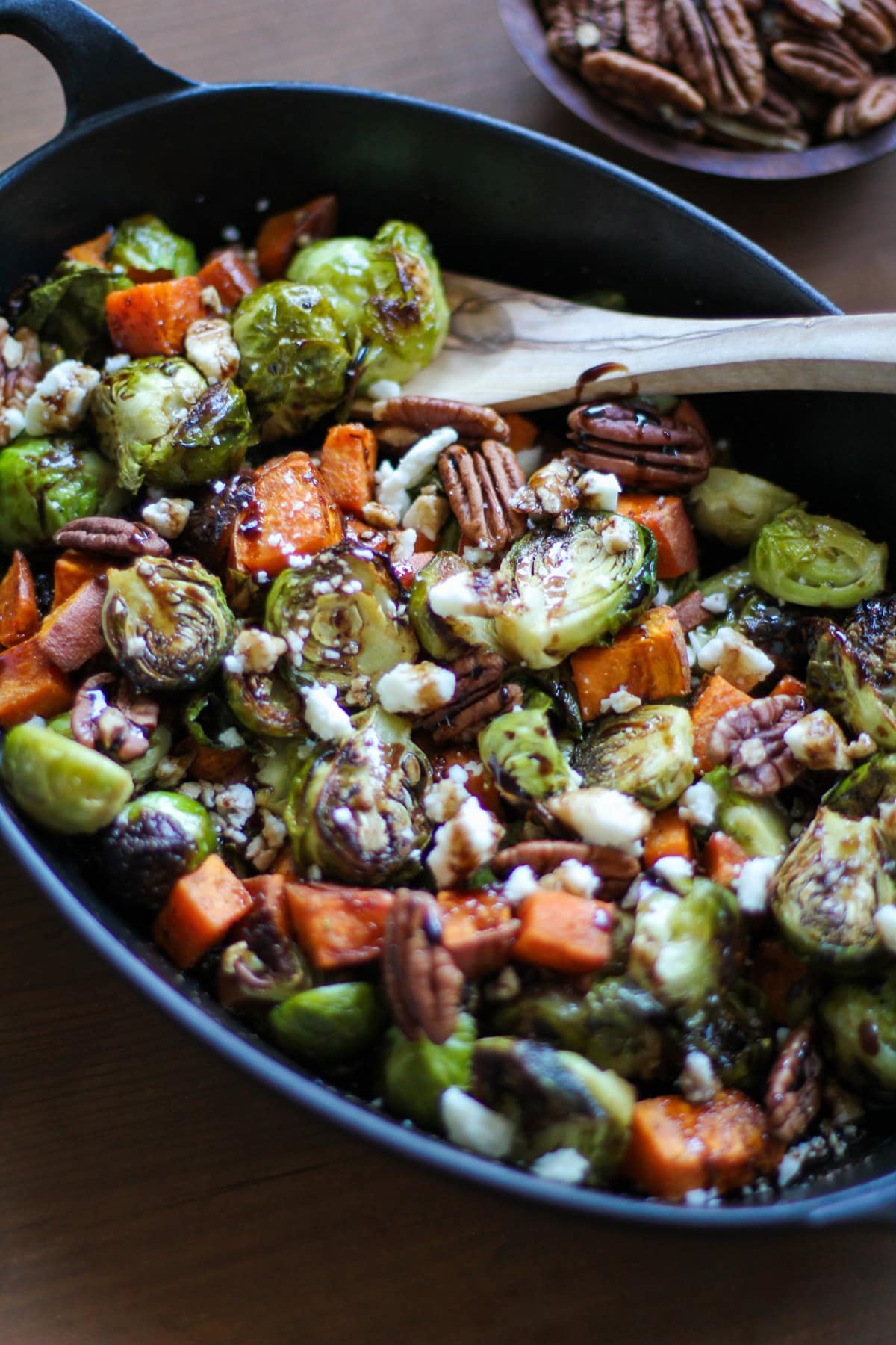 A cast iron skillet filled with roasted Brussels sprouts, sweet potato chunks, whole pecans, and crumbled cheese sits invitingly with a wooden spoon inside. A bowl of pecans is visible in the background.