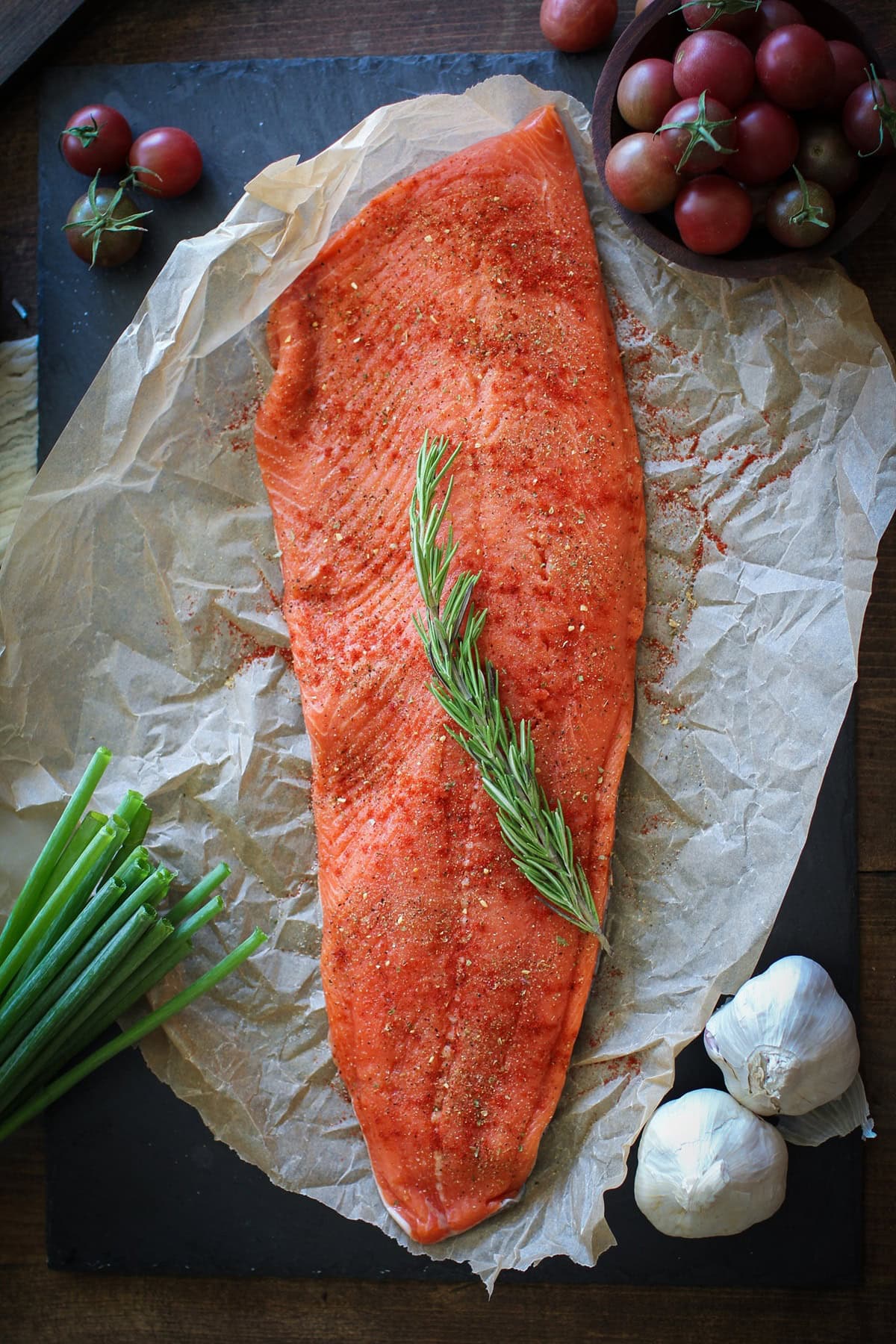 A large, raw salmon fillet prepared for roasted salmon, seasoned with spices and crowned with rosemary, rests on parchment paper. Green onions, garlic bulbs, and a bowl of cherry tomatoes are artfully arranged nearby on a dark surface.