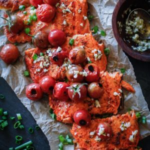 Long salmon filet on a black cutting board topped with cooked tomatoes, garlic, and chopped green onions, and cut into individual filets. Ready to serve.