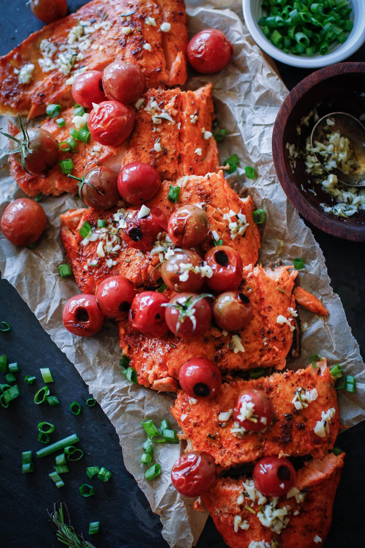 Long salmon filet on a black cutting board topped with cooked tomatoes, garlic, and chopped green onions, and cut into individual filets. Ready to serve.