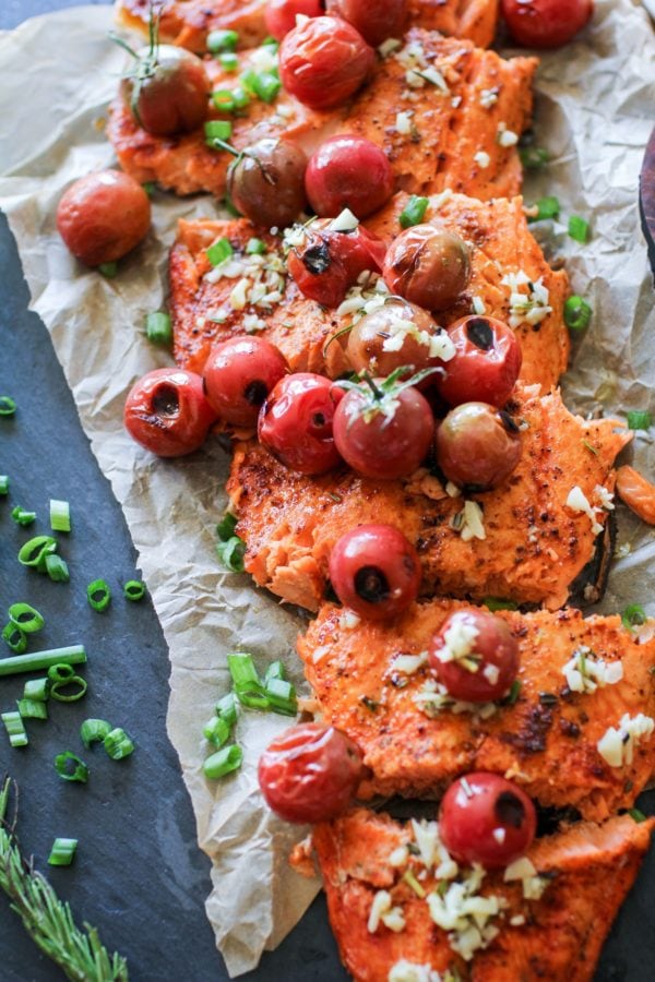Large salmon filet cut into individual filets on a large black cutting board with fresh garlic and tomatoes on top. Fresh rosemary and chopped green onions to the side, ready to serve.
