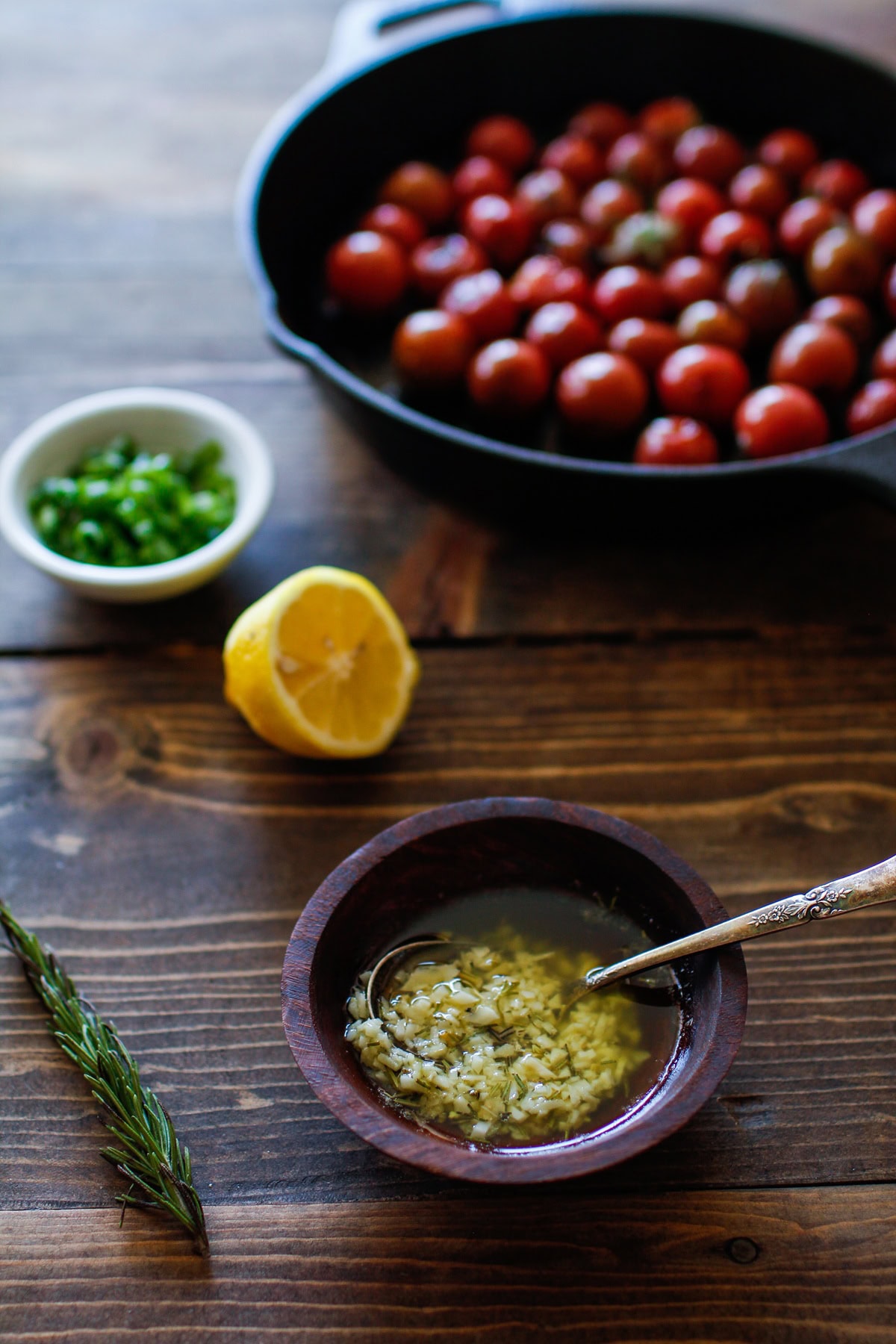 Small bowl of melted garlic butter and rosemary with fresh rosemary to the side, a skillet of tomatoes and a lemon in the background.