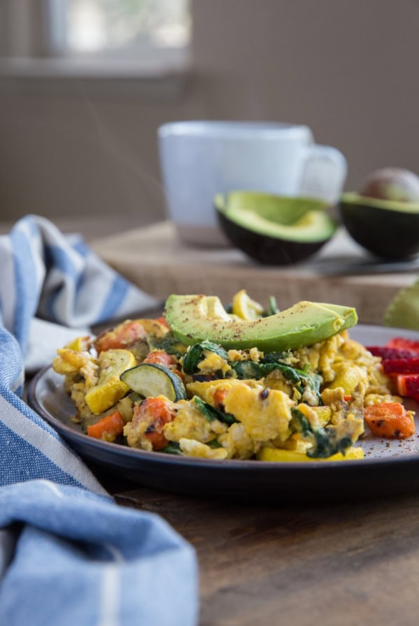 Plate of roasted vegetable scramble steaming with a mug of coffee or tea in the background. Sliced avocado on top of the scramble and sliced strawberries on the side of the plate.