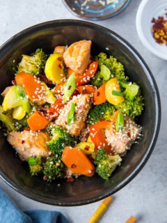 Black bowl of stir fry salmon and vegetables with chopsticks to the side and a dark blue napkin, ready to consume.