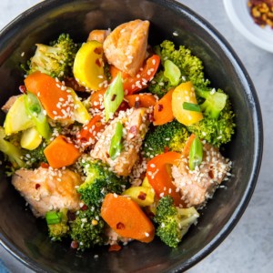 Black bowl of stir fry salmon and vegetables with chopsticks to the side and a dark blue napkin, ready to consume.