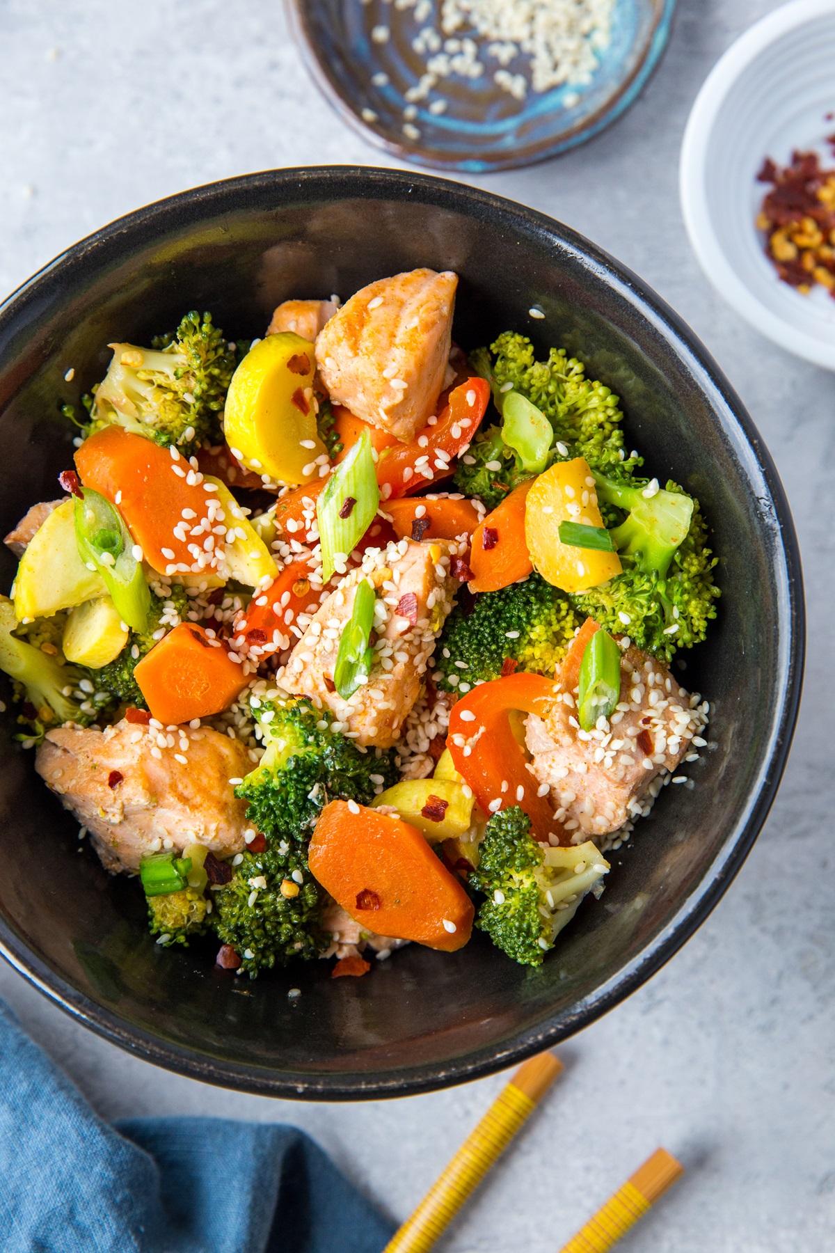 Black bowl of stir fry salmon and vegetables with chopsticks to the side and a dark blue napkin, ready to consume.