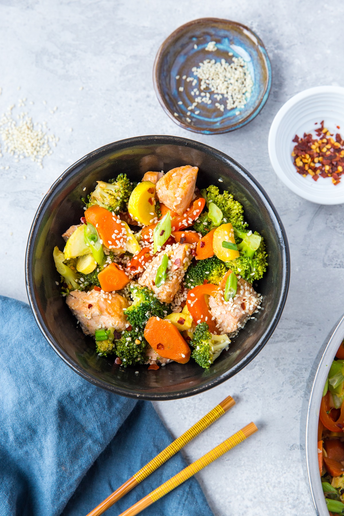Black bowl of salmon stir fry and vegetables with chop sticks and a blue napkin to the side.