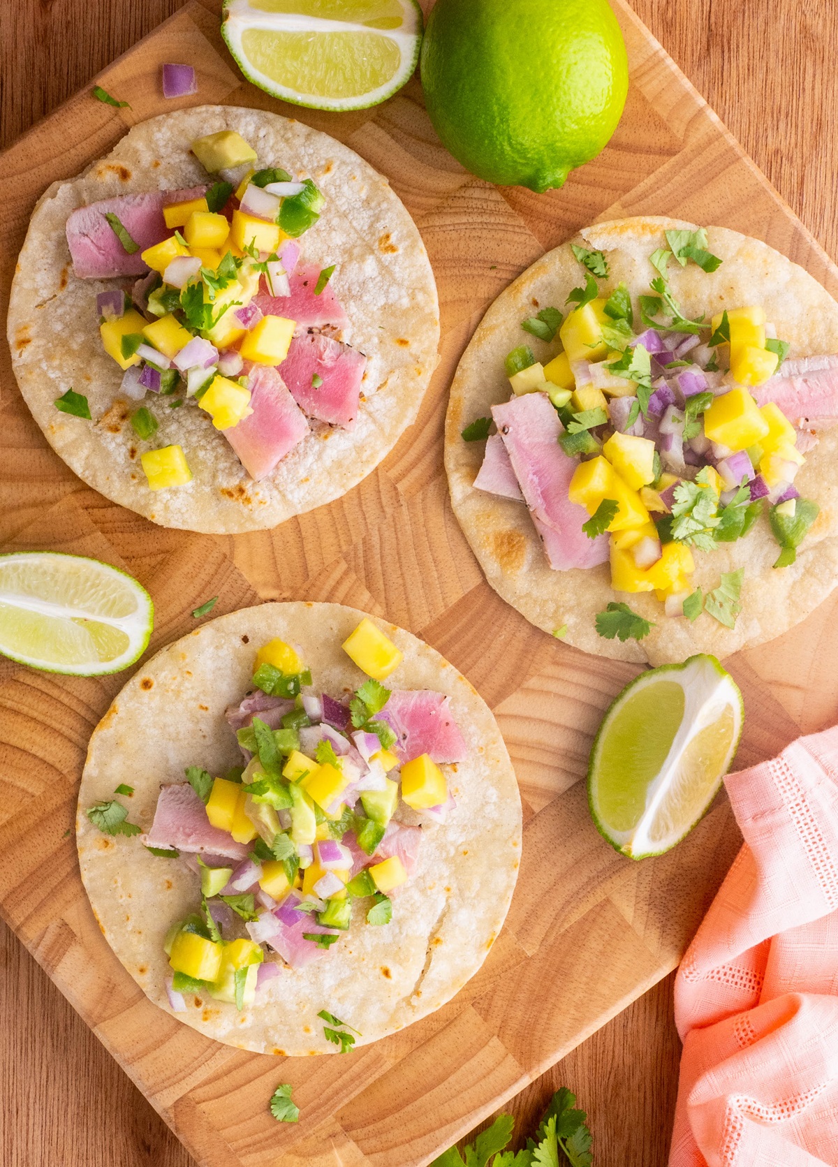 Three Seared Ahi Tacos with Mango Salsa on a wooden cutting board with fresh cilantro and limes to the side.