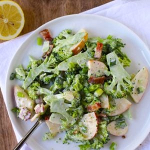 Shaved broccoli on a plate with bacon, apples and a glass of wine to the side. A napkin underneath the plate of salad.