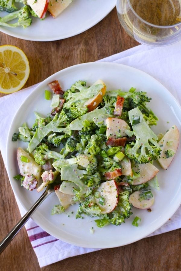 Shaved broccoli on a plate with bacon, apples and a glass of wine to the side. A napkin underneath the plate of salad.
