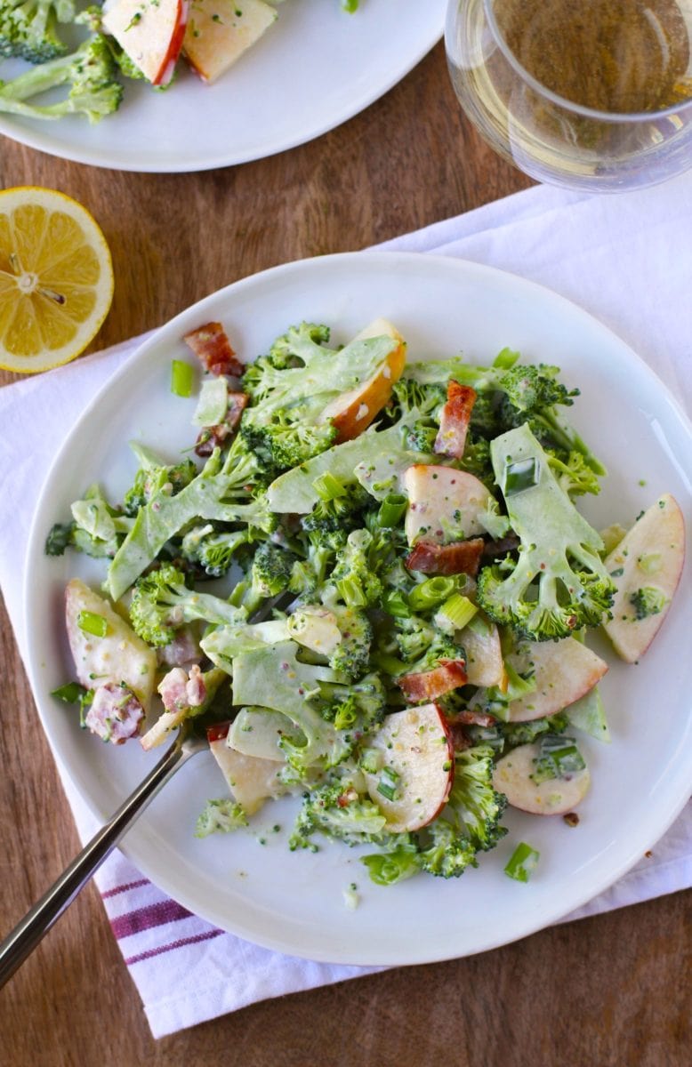Shaved broccoli on a plate with bacon, apples and a glass of wine to the side. A napkin underneath the plate of salad.
