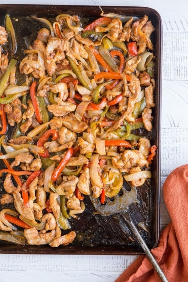 Sheet pan with chicken fajitas on top with a metal spatula scooping some off and a red napkin to the side.