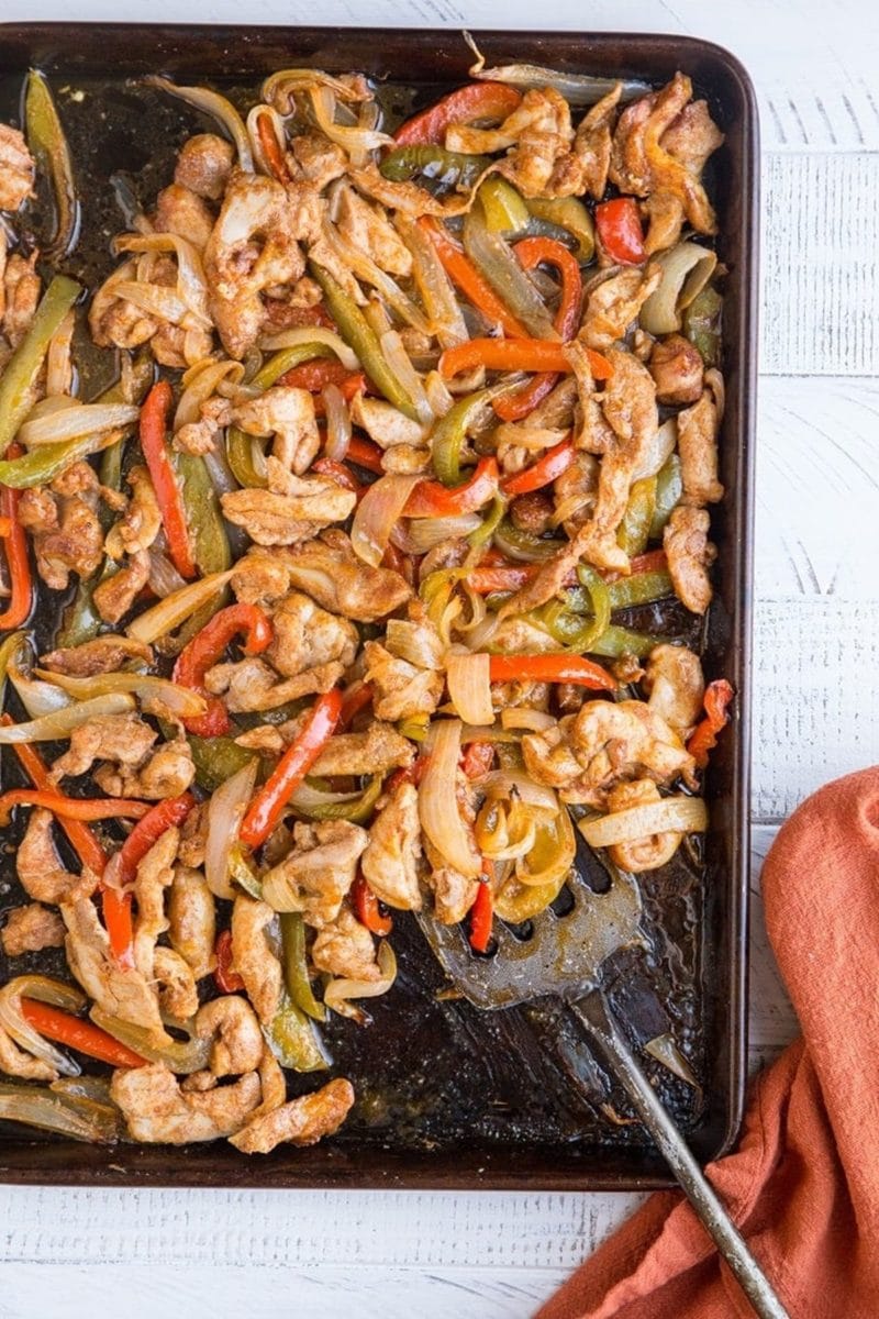 Sheet pan with chicken fajitas on top with a metal spatula scooping some off and a red napkin to the side.