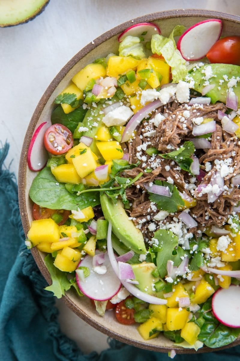 Shredded Beef Taco Salad with mango salsa, queso fresco, avocado and cilantro-lime dressing in a bowl with fresh veggies and a blue napkin to the side.