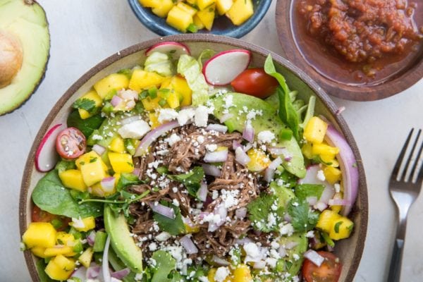 A colorful Taco Salad bowl with shredded beef, avocado slices, cherry tomatoes, radishes, mango chunks, spinach, onions, and cheese. Nearby are bowls of diced mango and salsa, plus a fork and half an avocado.