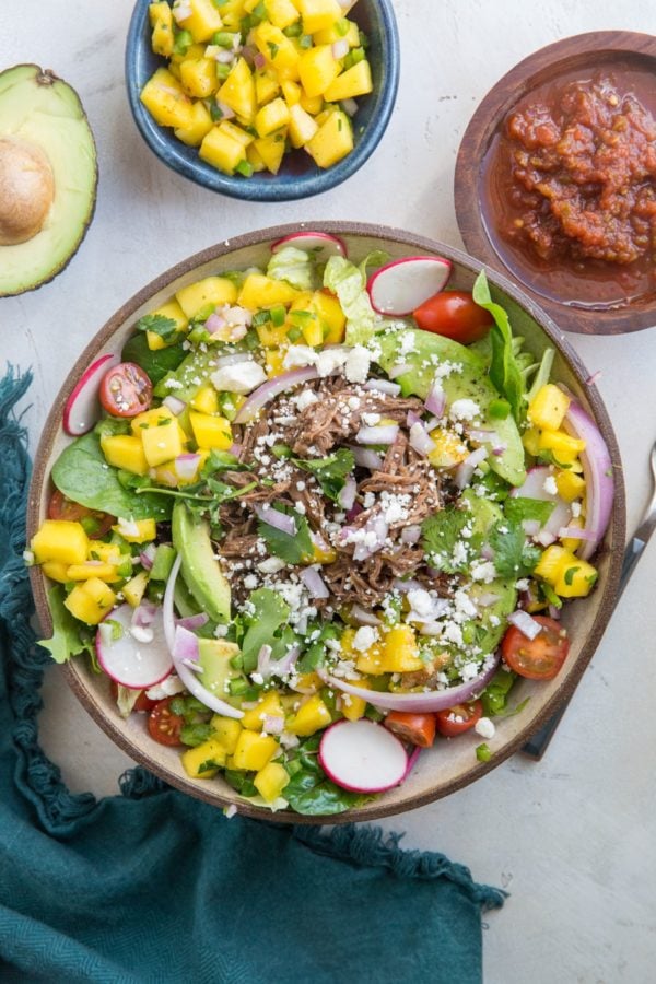 Large bowl of Shredded Beef Taco Salad with spring greens, avocado, cherry tomatoes, mango salsa, and more. A blue napkin to the side and a bowl of salsa.