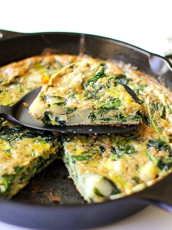 Cast iron skillet full of a frittata with spinach, leeps, and potatoes. Ready to serve with a slice taken out.